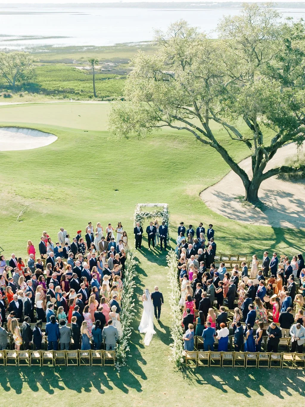 Bride walking down the flower-lined aisle during an outdoor wedding ceremony at the River Course on Kiawah Island.