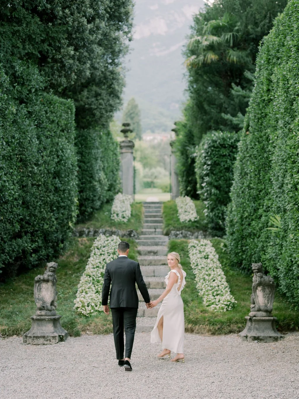 Couple walking through a formal villa garden in Italy with stone steps, manicured hedges, and white floral arrangements.