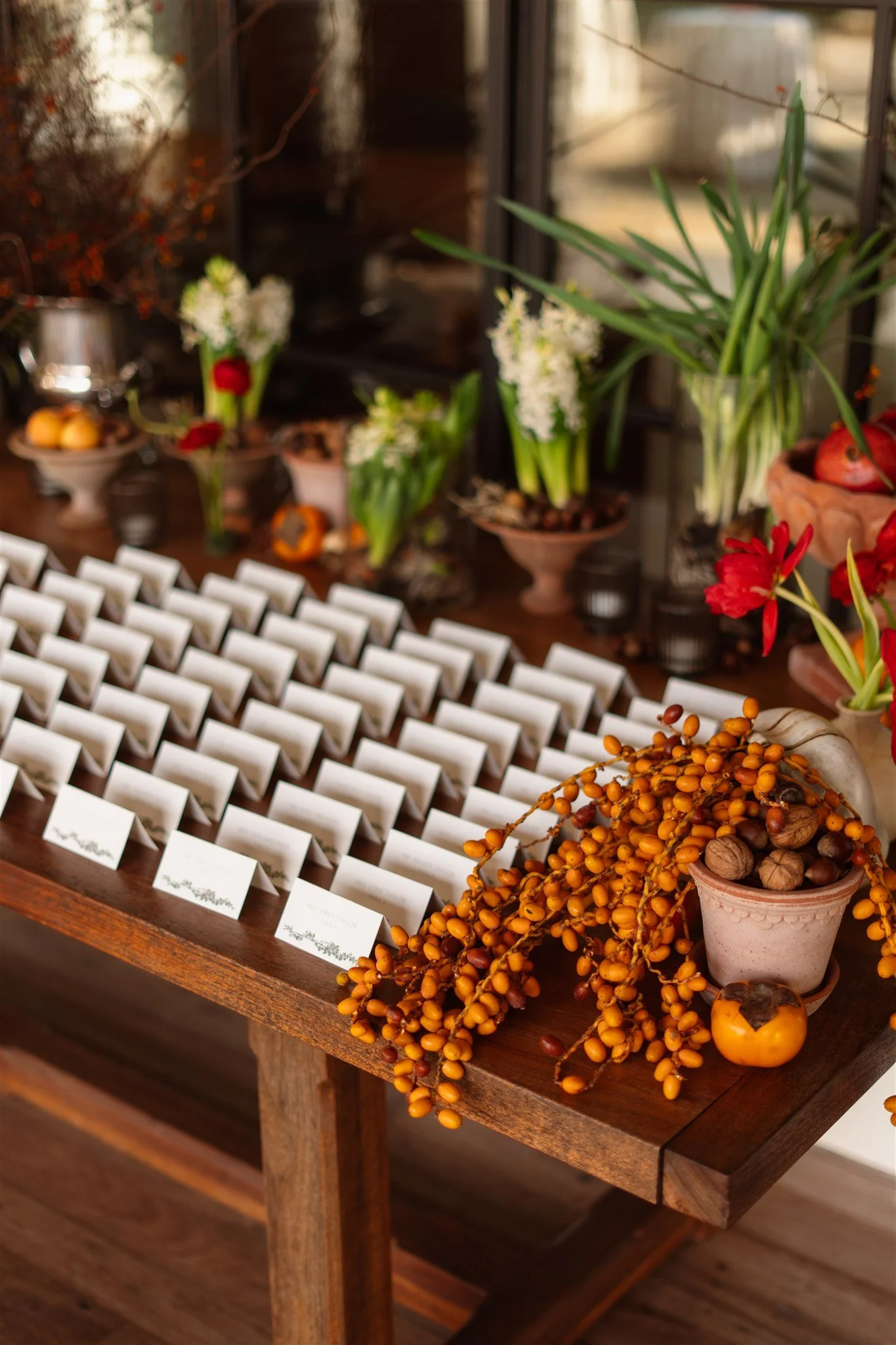 Escort card display on a wooden table with folded place cards, potted florals, citrus, walnuts, and orange berries for a destination wedding.