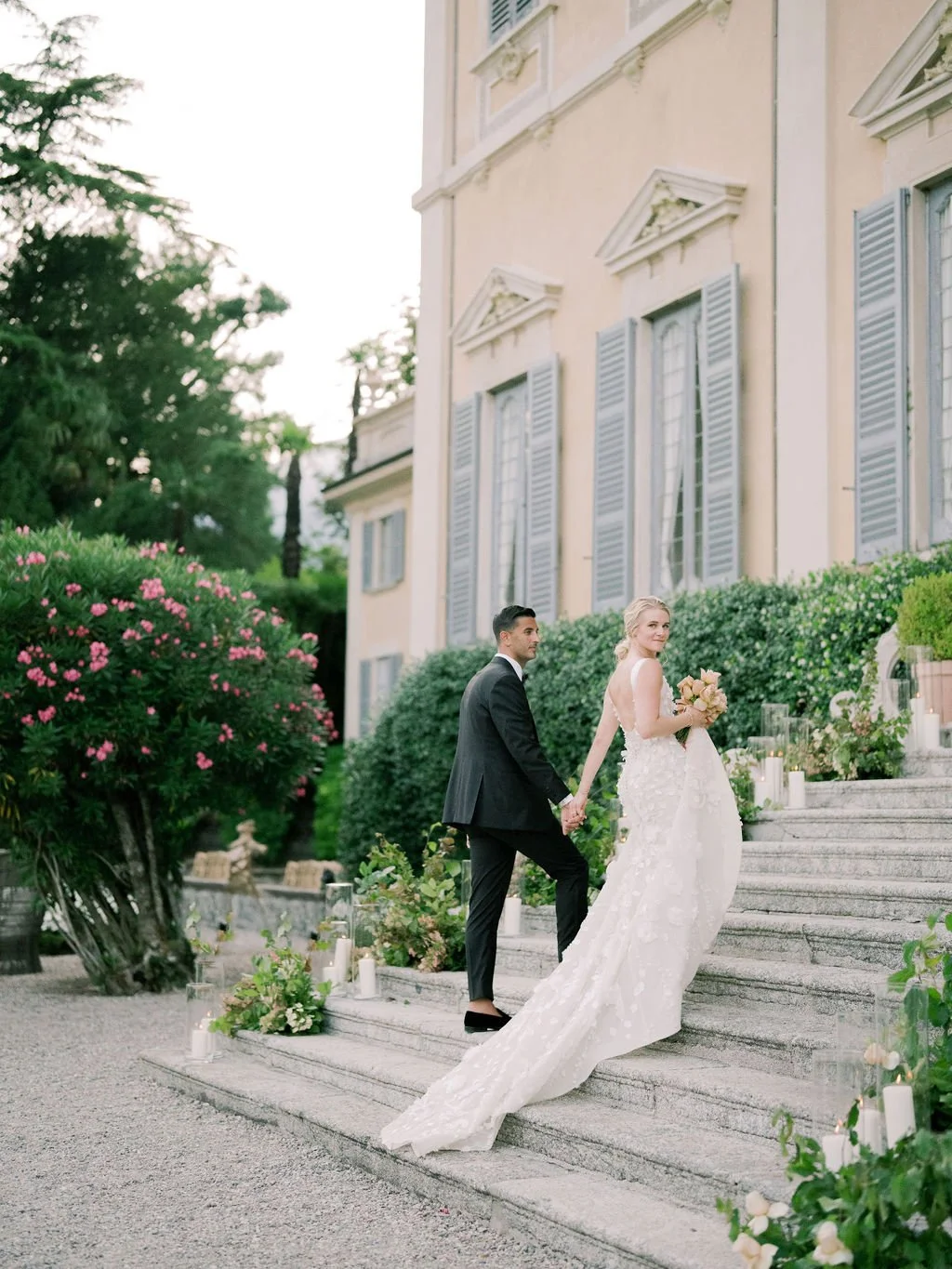 Bride and groom climbing the stone steps of a Lake Como villa decorated with soft florals, greenery, and glowing pillar candles.