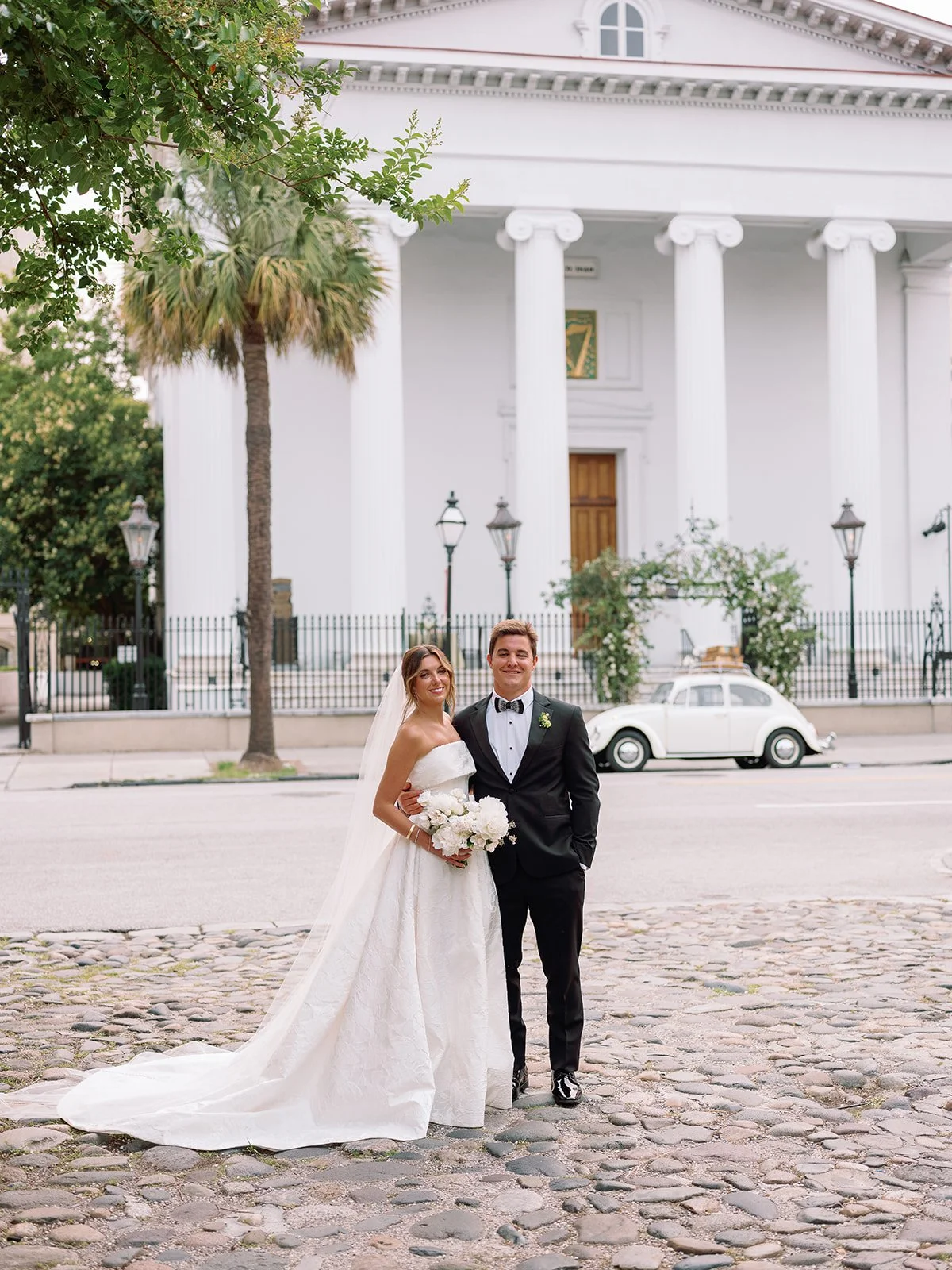 Bride and groom portrait on a cobblestone street with a historic Charleston church and vintage white car in the background.