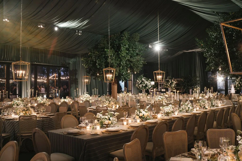 Candlelit tented wedding reception with green draping, hanging lanterns, and layered dinner tables.
