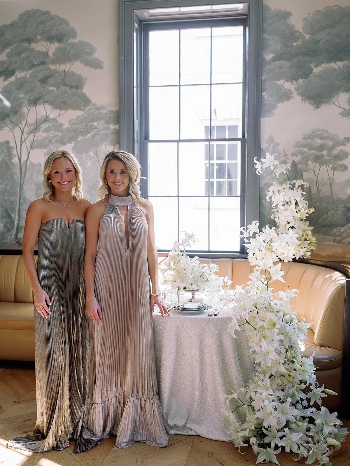 Two wedding planners stand beside a refined reception table with white lilies, patterned china, and soft natural light.