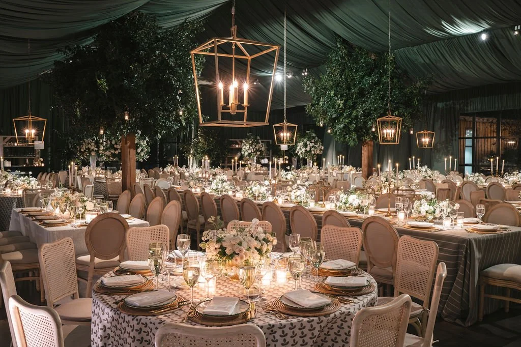 Candlelit wedding reception with deep green draping, suspended lanterns, layered dinner tables, neutral linens, and white floral arrangements.