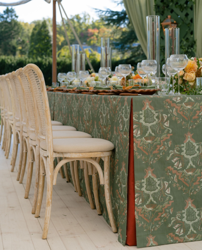 Long outdoor reception table with cane-back chairs, a green patterned tablecloth with rust accent panel, clear glassware, and tall cylinder candles.