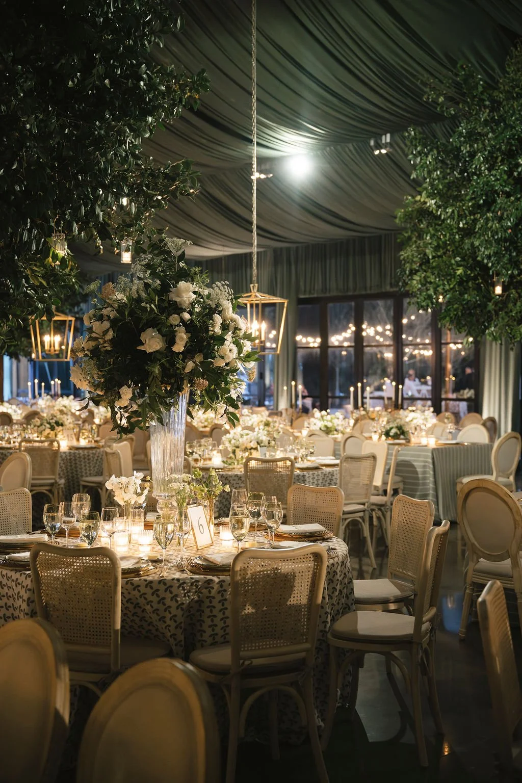 Candlelit tented wedding reception with tall white floral centerpieces, patterned linens, and green ceiling draping.