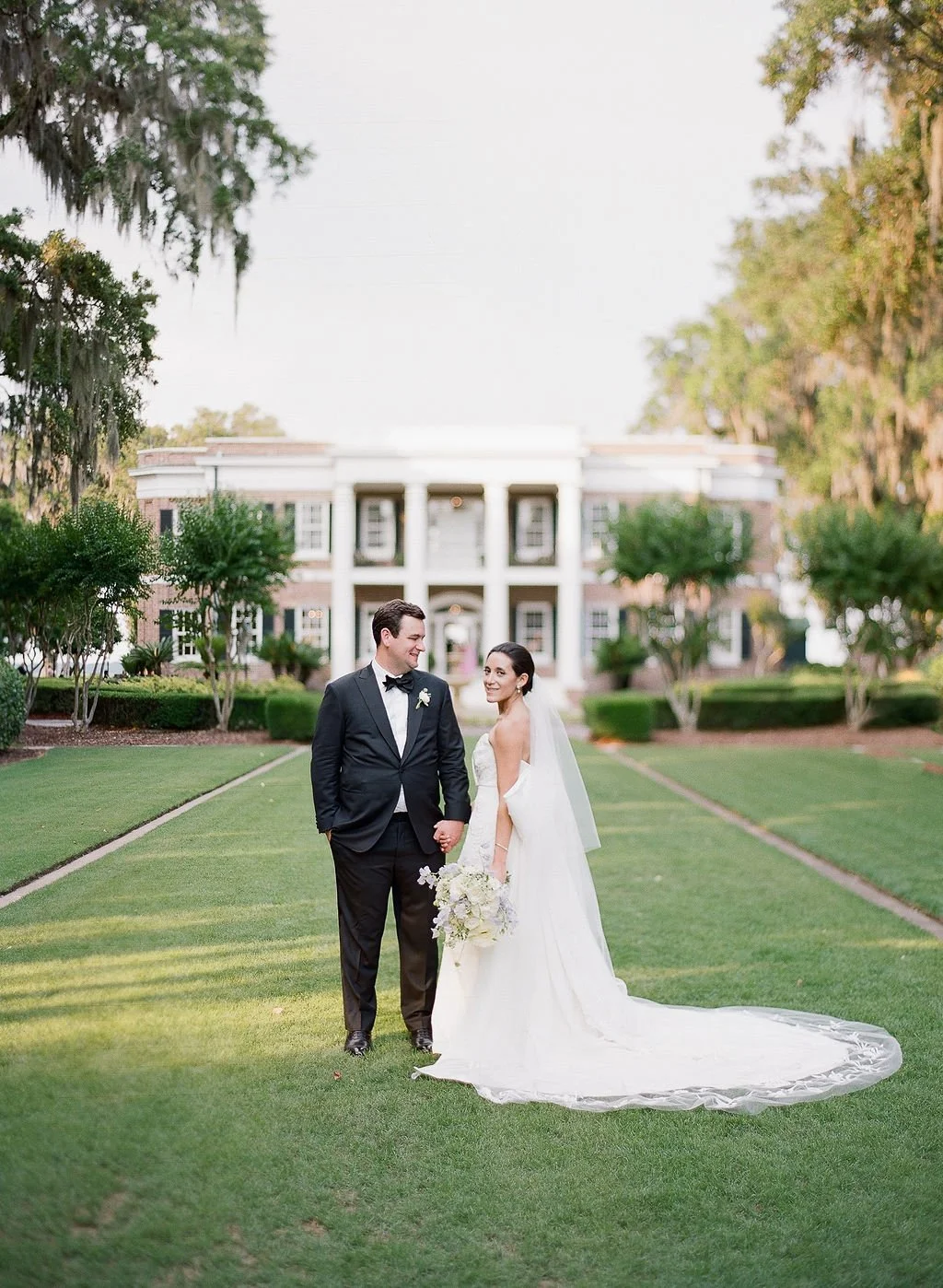 Bride and groom standing on the front lawn of a historic Southern estate wedding venue framed by oak trees and white columns.