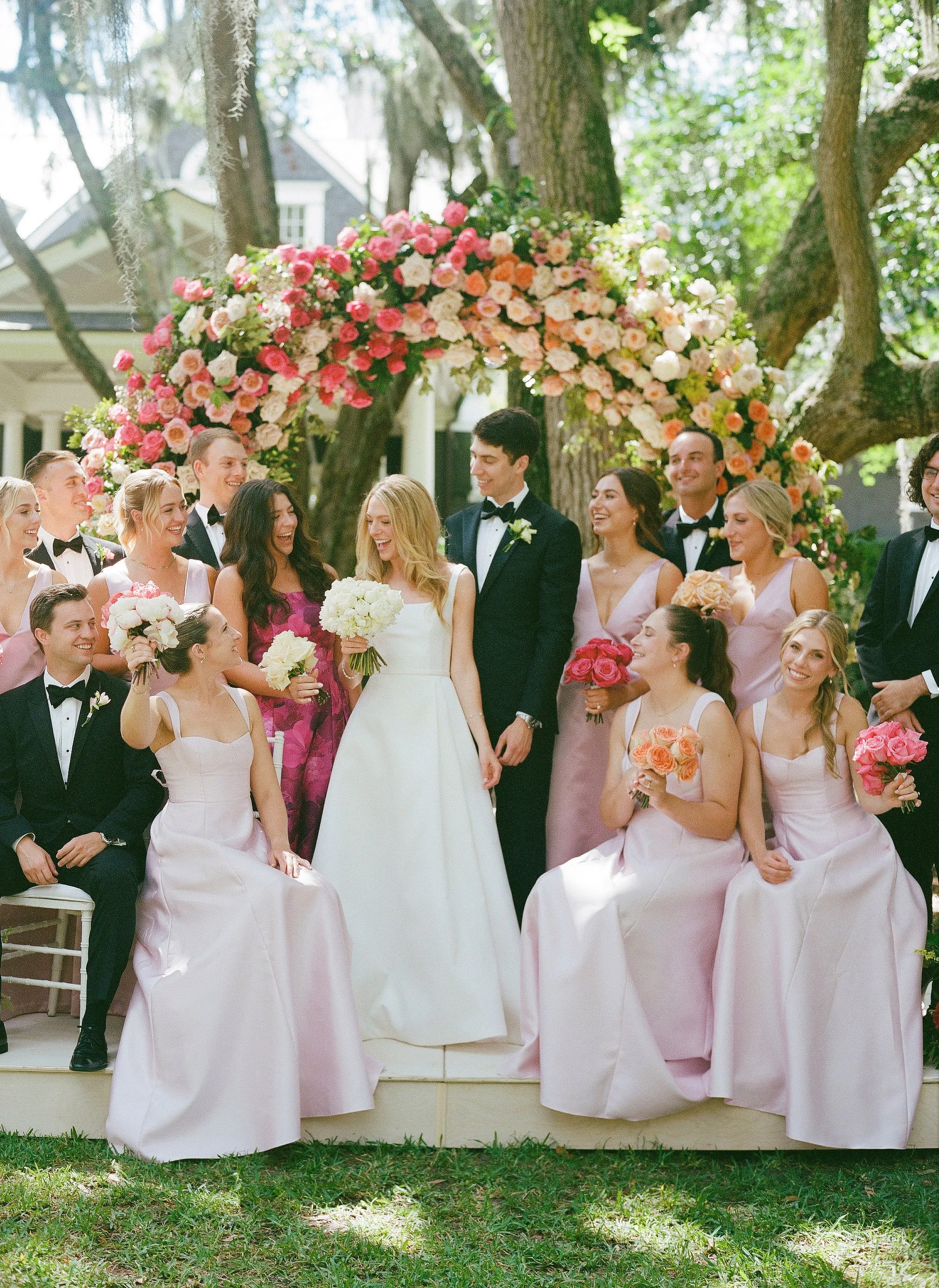 Wedding party gathered beneath a lush floral arch in pink and coral tones under live oaks, with the bride in a classic white gown and attendants in blush dresses.