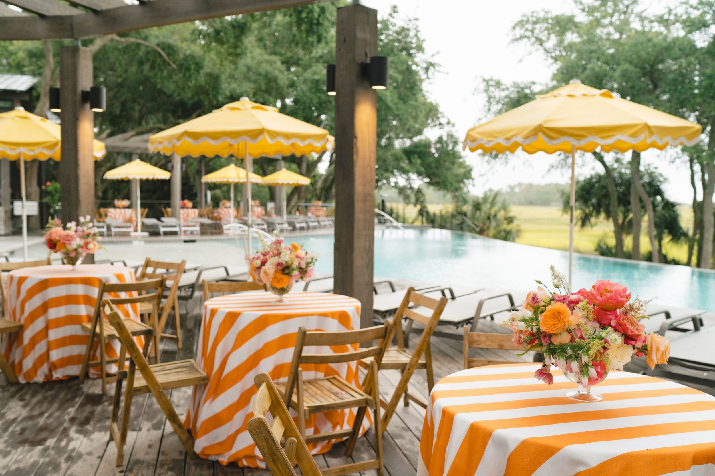 Poolside welcome gathering with yellow umbrellas, orange-and-white striped table linens, and bright floral arrangements overlooking the marsh on Kiawah Island.