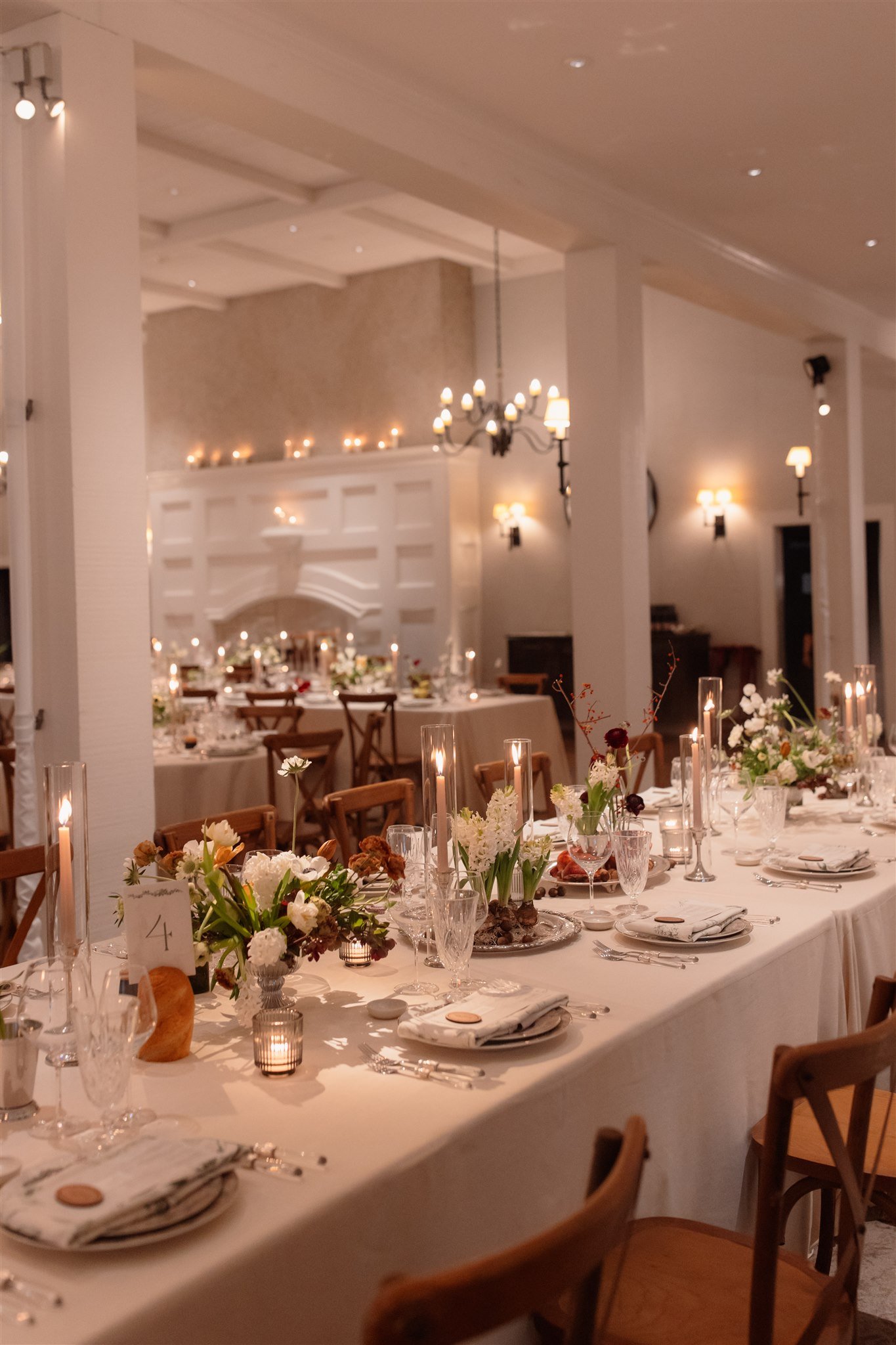 Indoor reception dinner with a long linen-draped table, crossback chairs, low florals, and tall glass candle hurricanes in a classic white room.