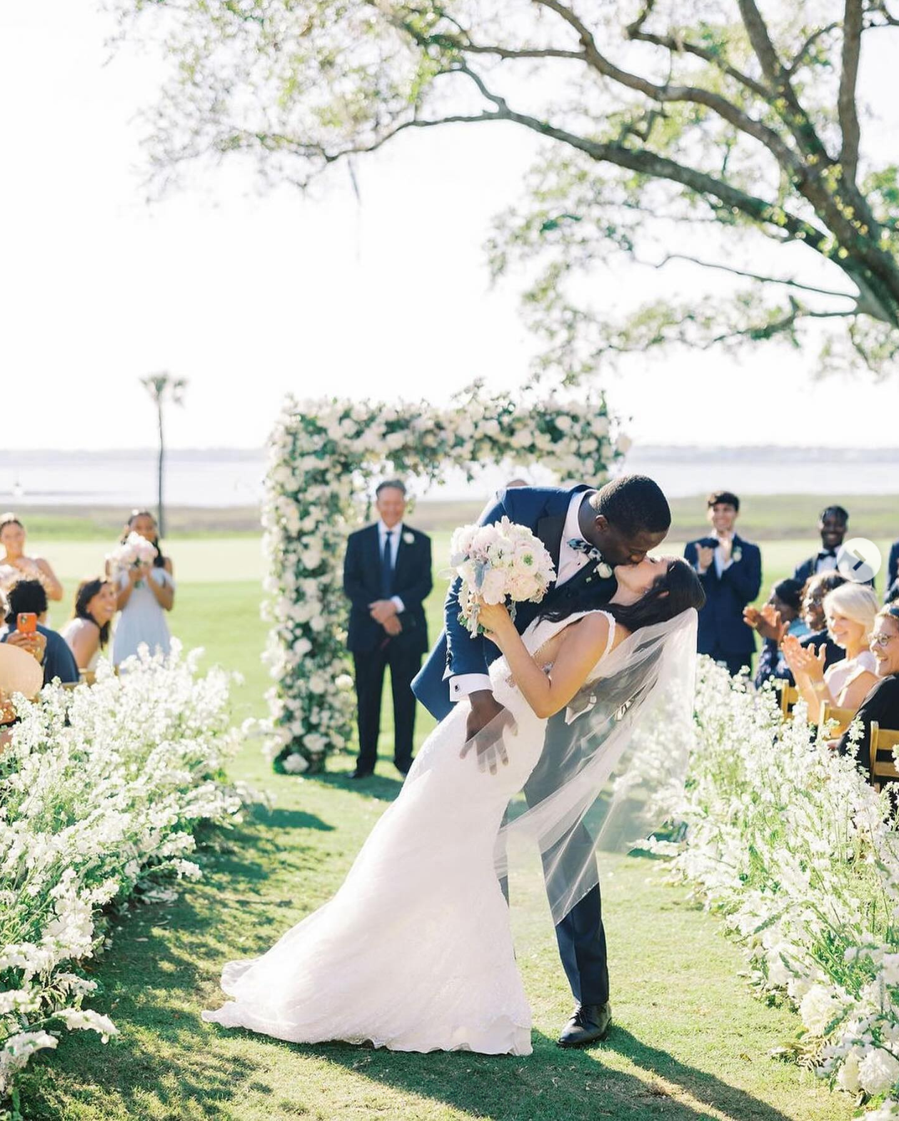Newlyweds share a kiss on a grassy ceremony aisle lined with white florals, with a floral altar arrangement and waterfront views in the background.