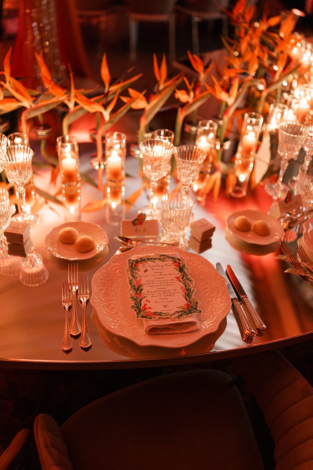 Candlelit wedding tablescape with bird of paradise florals, crystal glassware, personalized menus, and layered place settings.