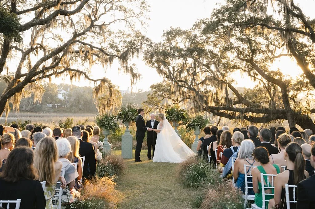 Outdoor Lowcountry wedding ceremony beneath oak trees with Spanish moss, waterfront views, and guests seated along the aisle.