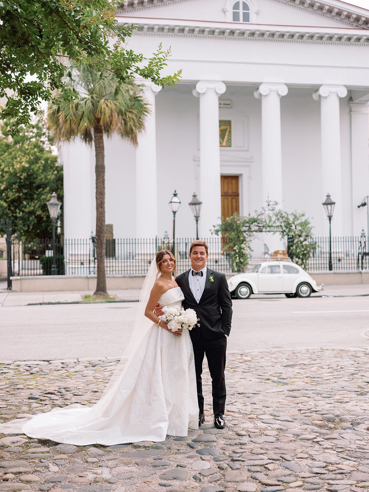 Bride and groom portrait on a cobblestone street in historic Charleston with a church and vintage car behind them.
