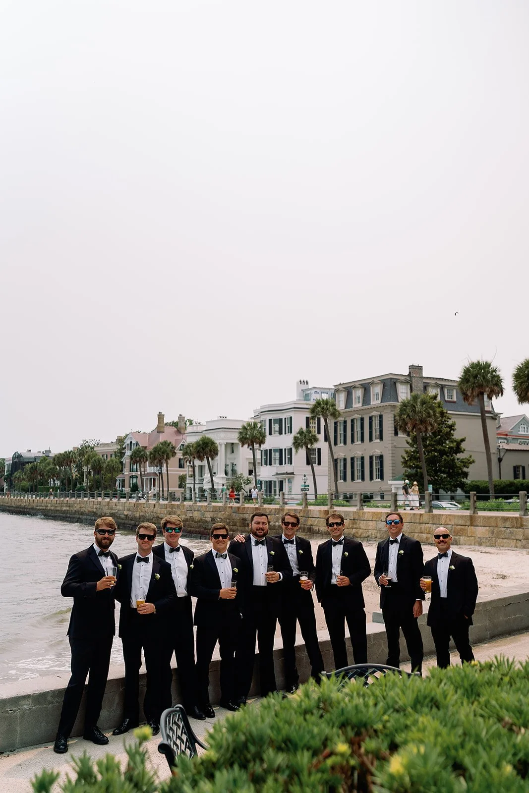 Alt text: Groom and groomsmen in black tuxedos gathered along the Charleston waterfront with historic homes and palm trees in the background.