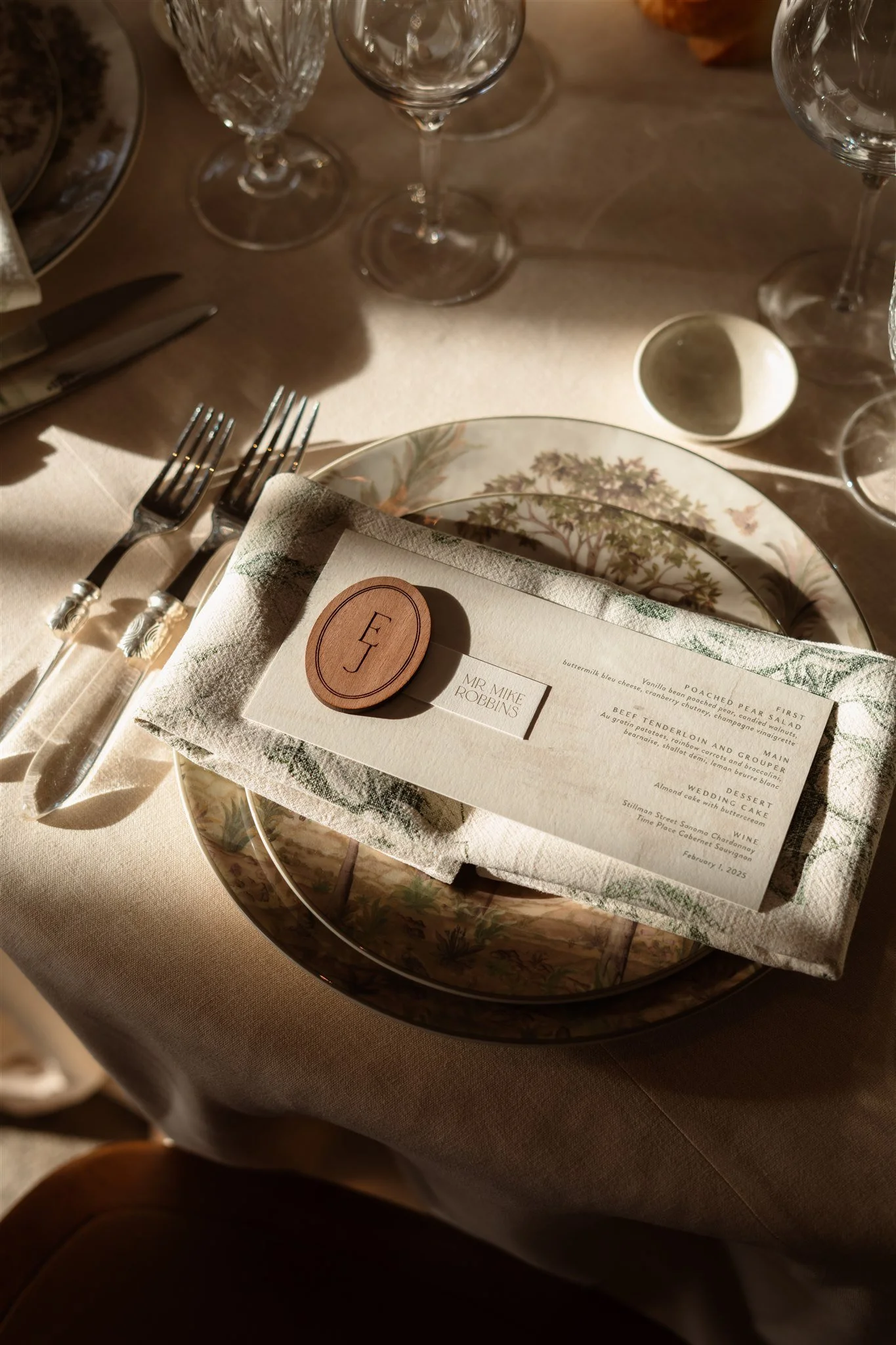 Close-up of a wedding place setting with botanical china, linen napkin, printed menu, and a wooden monogram accent in warm light.