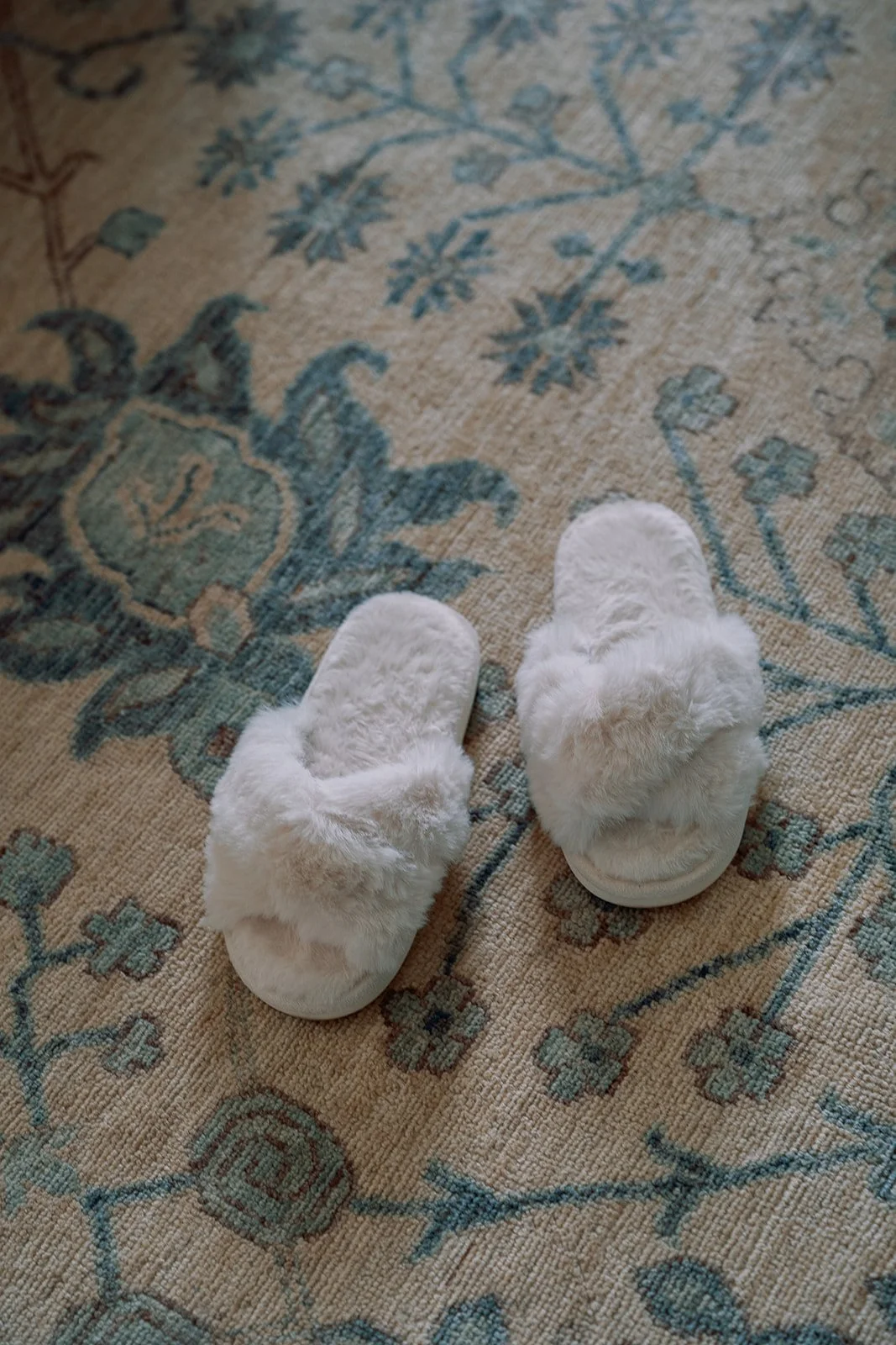 Alt text: White plush slippers resting on a blue and neutral patterned rug during wedding day preparations in Charleston.