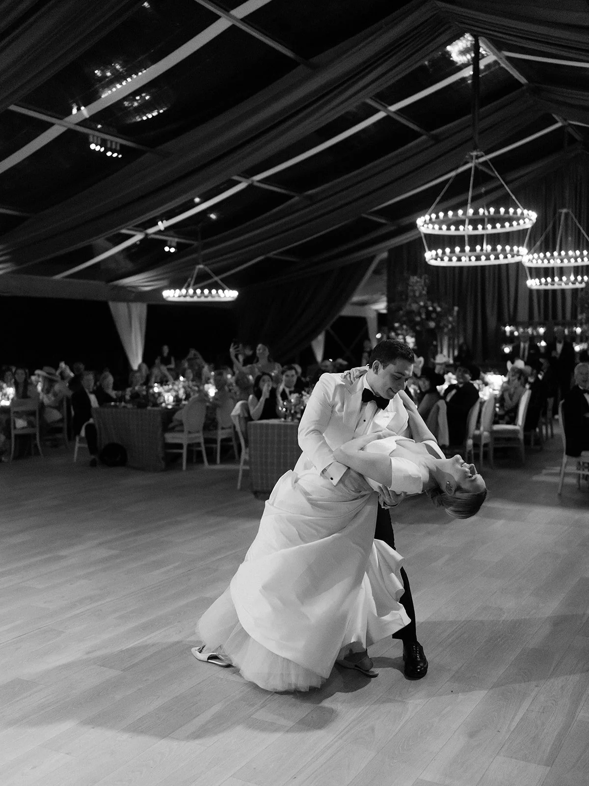 Bride and groom dancing beneath circular chandeliers during an evening tented wedding reception surrounded by seated guests.