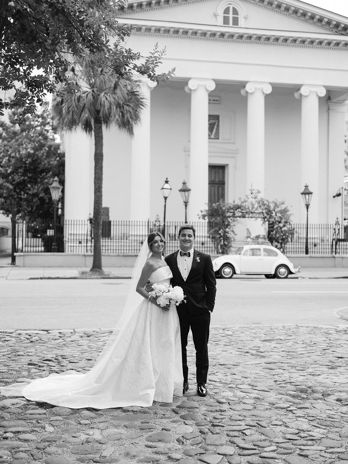 Black-and-white portrait of a bride and groom on a cobblestone street in front of a white columned building with a vintage car in the background.