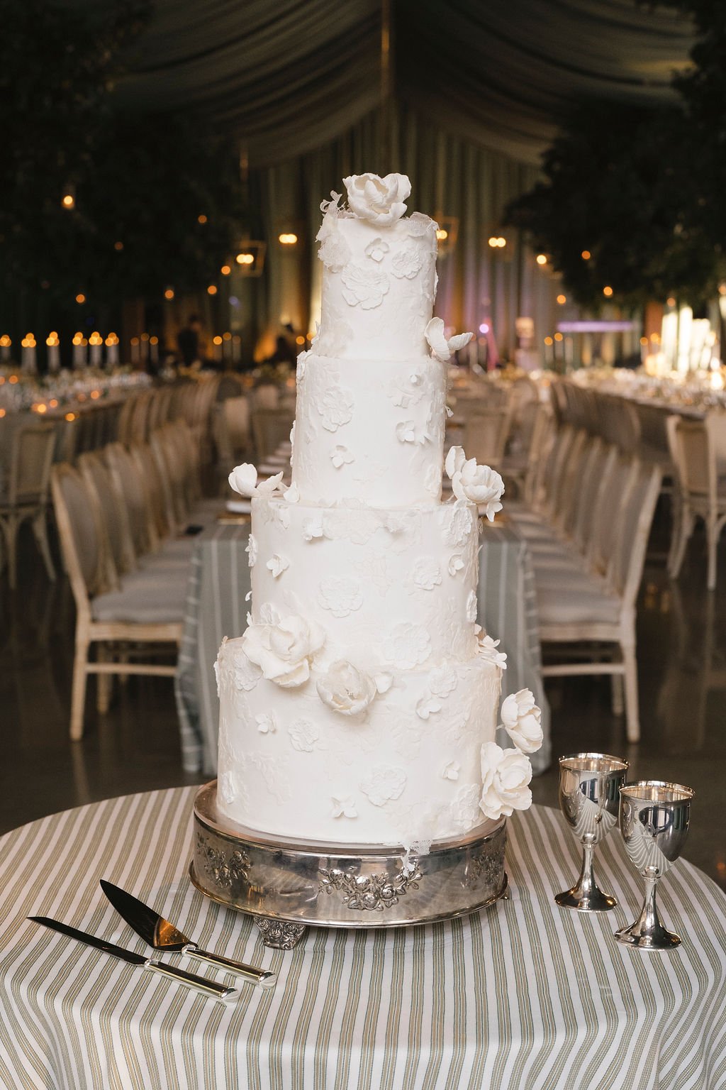 Four-tier white wedding cake adorned with delicate floral appliqués, displayed on a silver stand with striped linen and polished silver goblets at an elegant Charleston reception designed by Reagan Events.