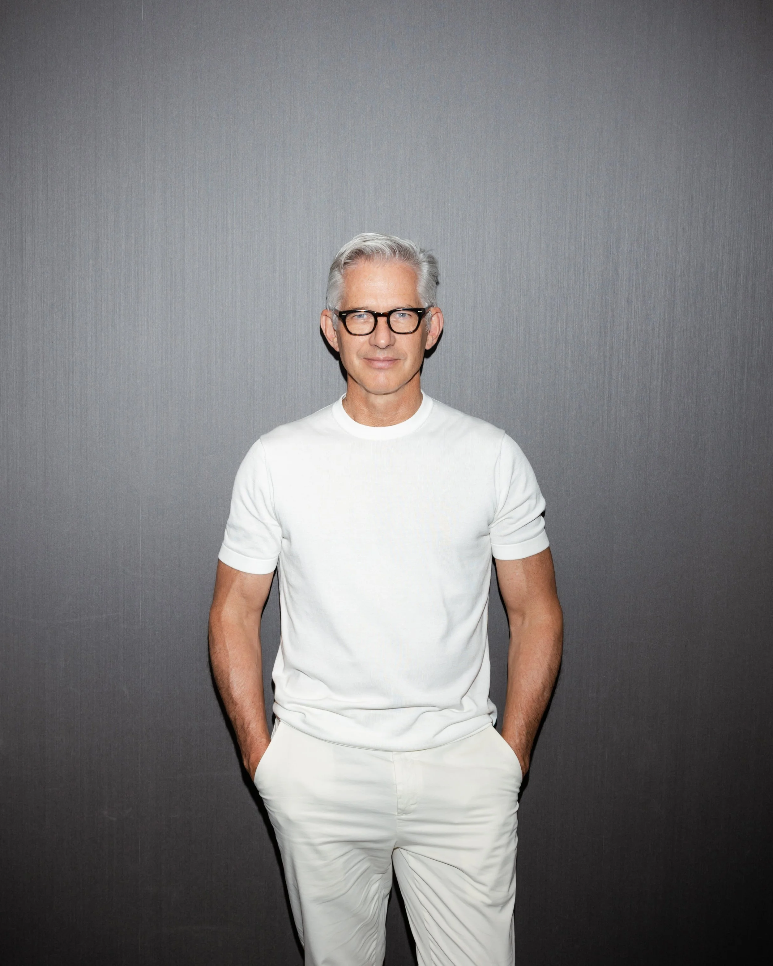 Bryan Rafanelli standing against a textured gray wall in a white shirt and light trousers for an editorial-style portrait.