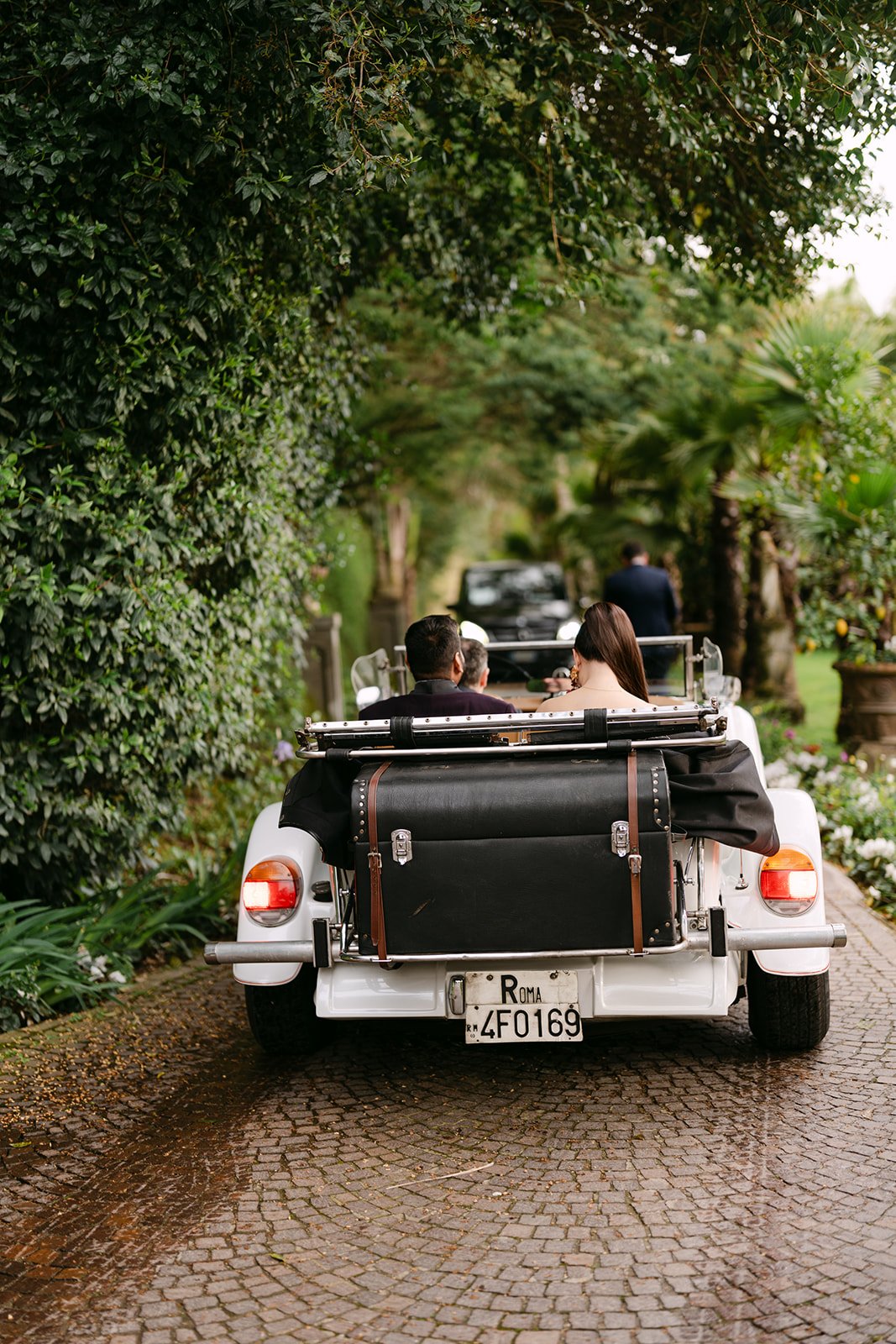 Couple riding through a lush garden in a vintage white convertible during a destination wedding celebration in Rome.