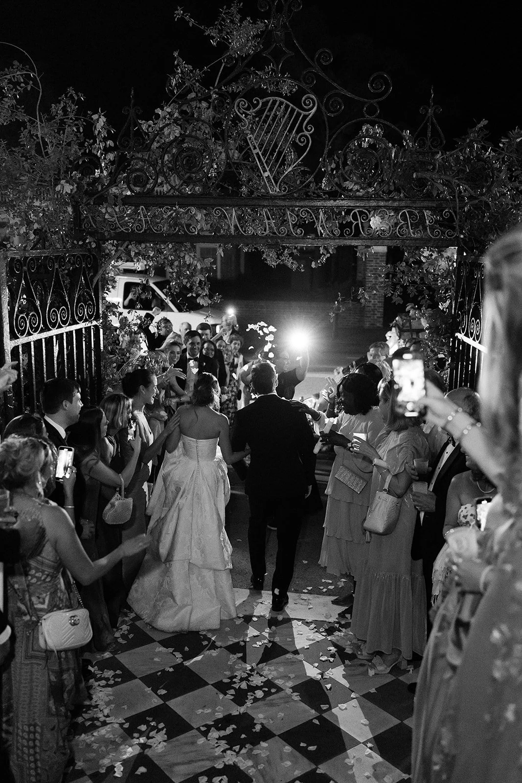 Black-and-white photo of a bride and groom exiting through an ornate iron gate at night as guests line the walkway and toss petals.