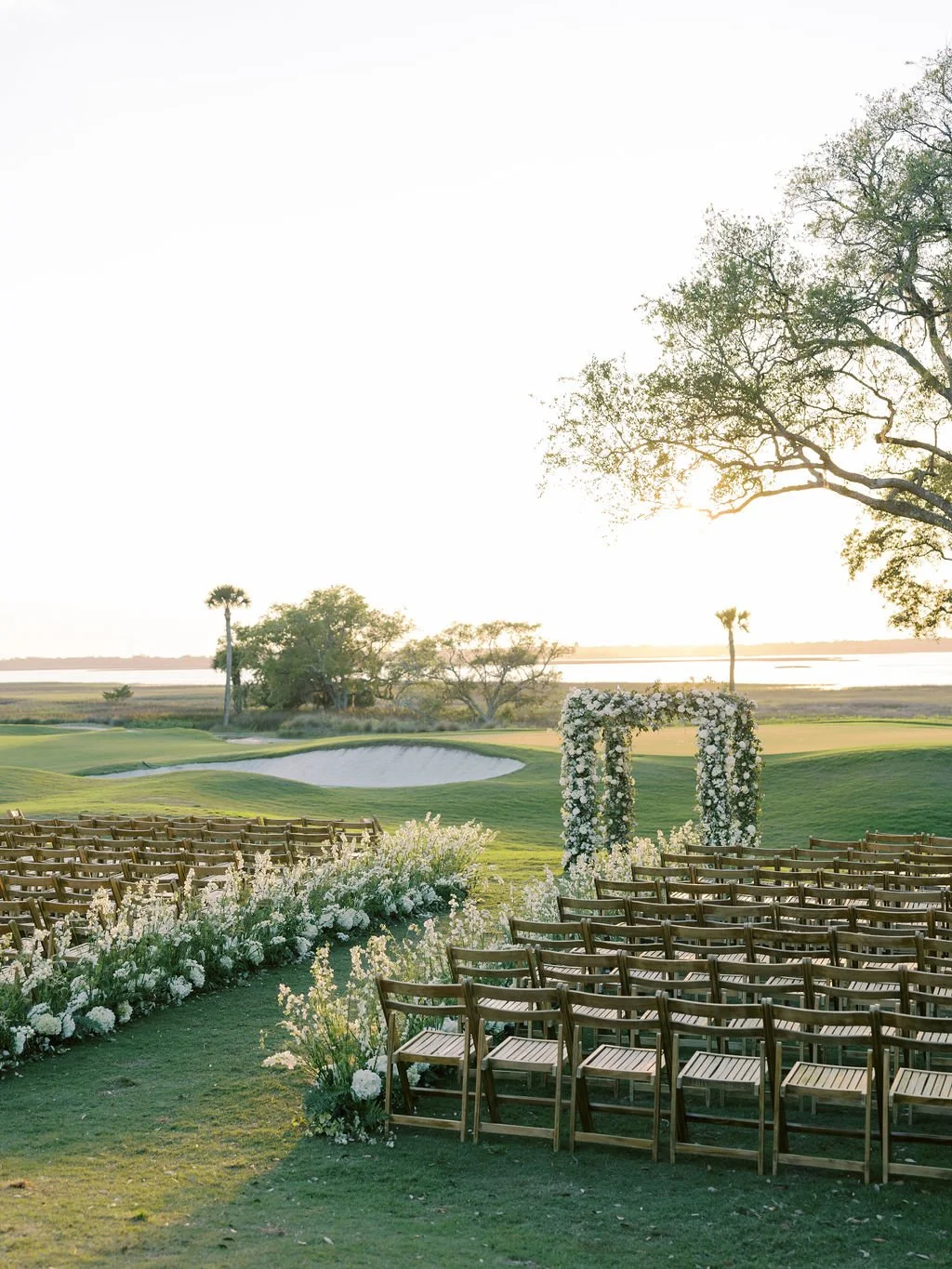 Outdoor Kiawah Island wedding ceremony setup with wooden chairs, white aisle flowers, a floral arch, and coastal marsh views at sunset.
