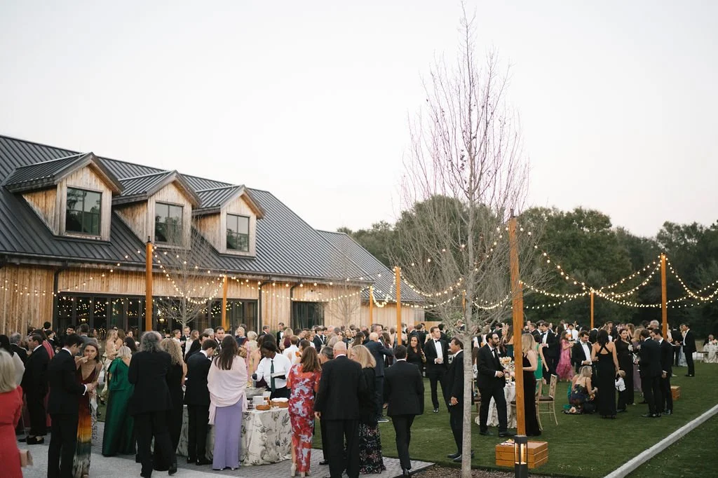 Guests gather for an outdoor wedding weekend cocktail hour under string lights beside a barn-style venue.