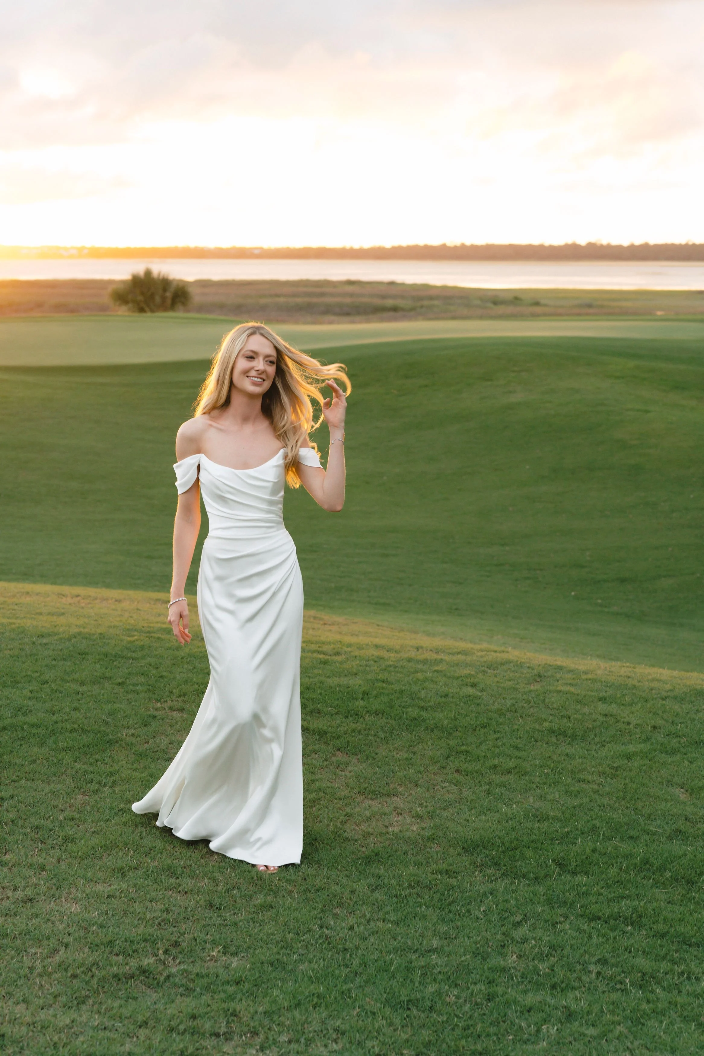 Bride in an off-the-shoulder satin gown walks across a green at sunset with marsh and water views, a quiet portrait during a Kiawah Island wedding weekend.