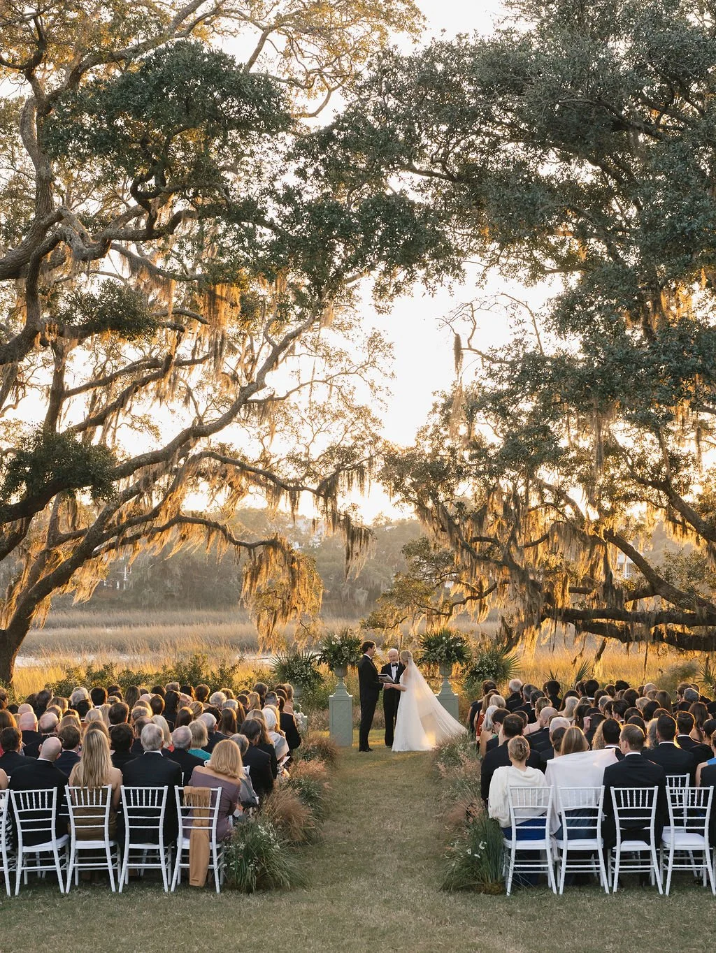 Outdoor wedding ceremony beneath sprawling live oak trees and Spanish moss, with guests seated along a grass aisle at sunset.