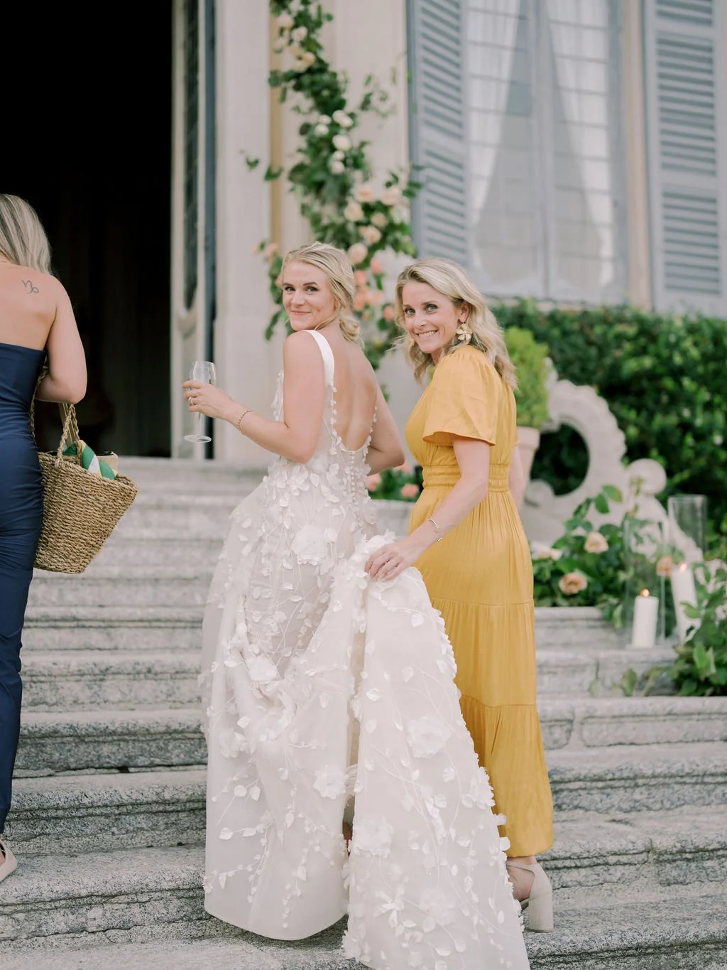 A bride ascending the stone villa steps alongside Reagan, framed by soft florals and shuttered windows during an Italy destination wedding celebration.