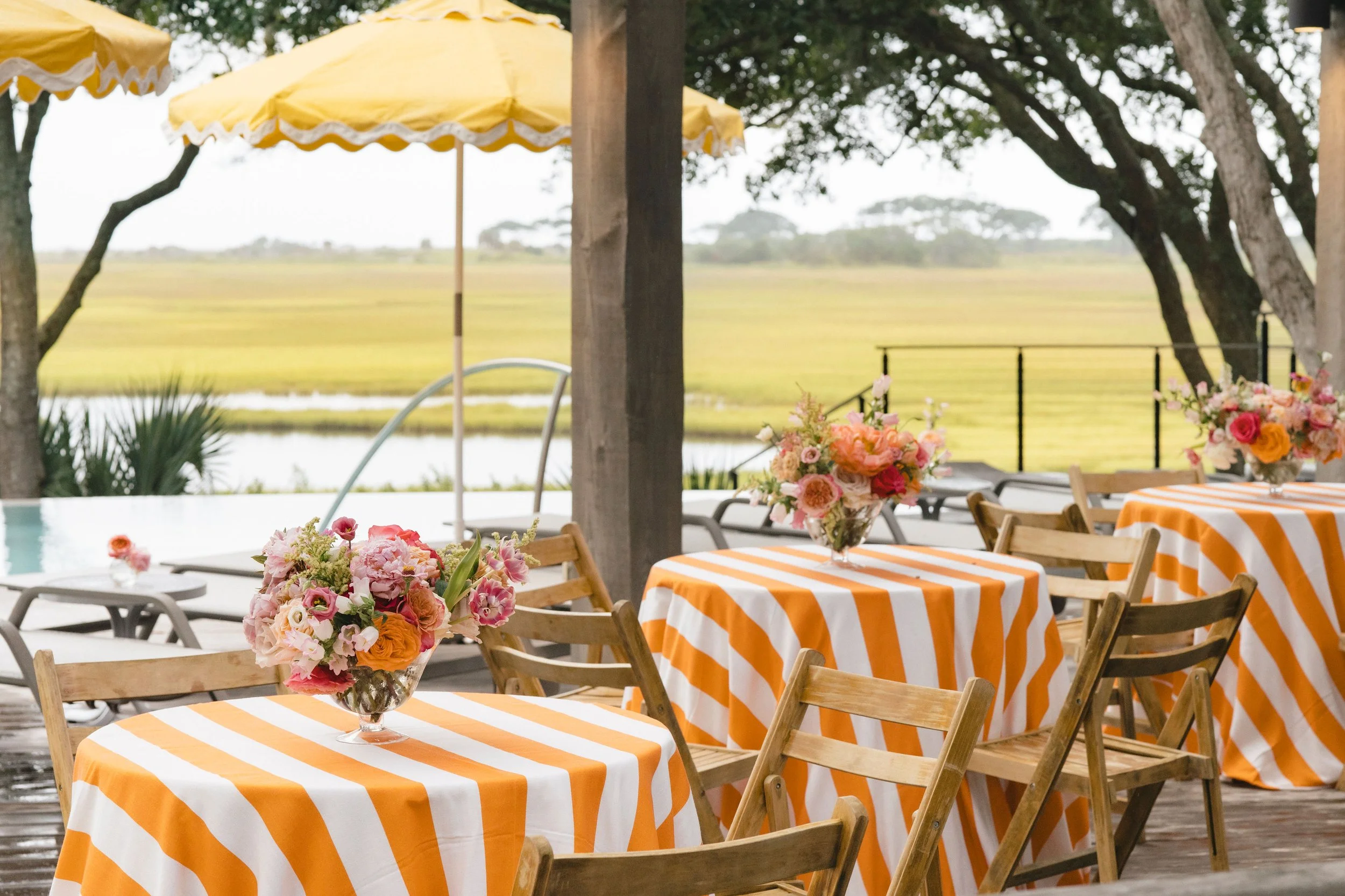 Close view of poolside brunch tables with orange striped linens, colorful florals, and marsh views for a wedding weekend brunch.
