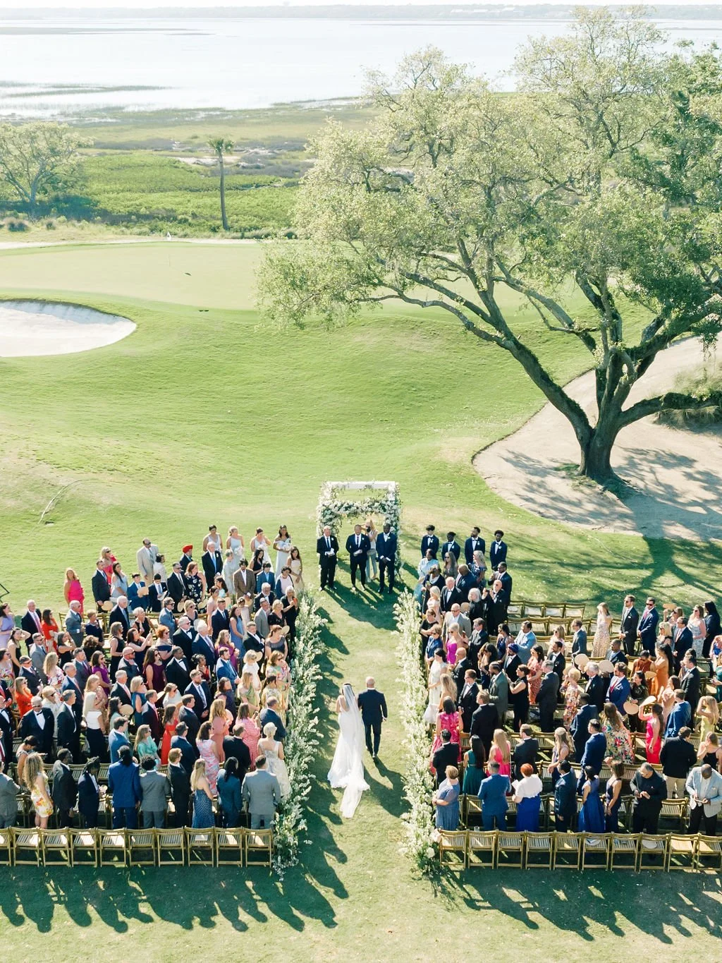 Overhead view of a large outdoor wedding ceremony on a coastal lawn with a long floral-lined aisle, guests seated on both sides, and marsh water in the distance.