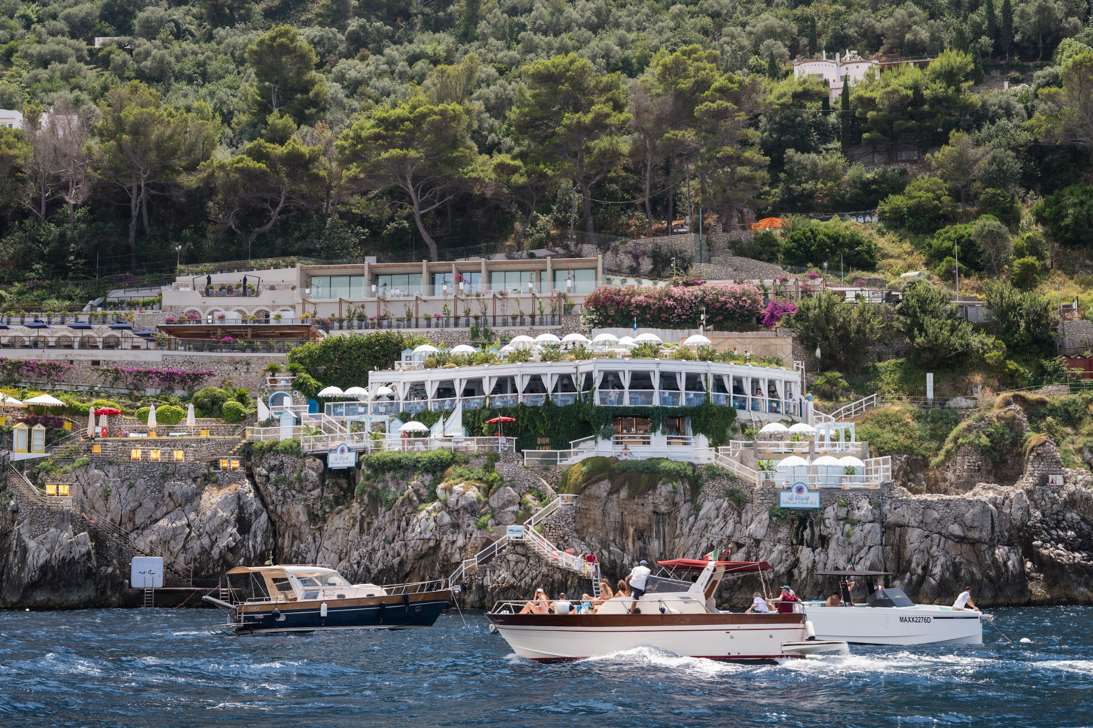 Boats cruising along the rocky Capri coastline beneath a cliffside restaurant and terraces.