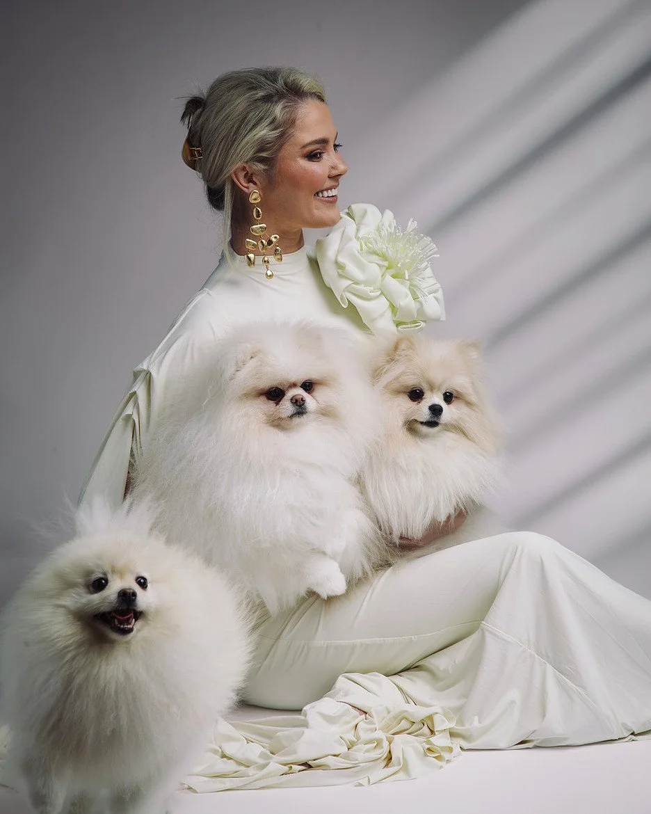 Podcast guest in a sculptural white outfit holding two small dogs, with a third dog in front, styled for an editorial portrait tied to a personal branding conversation.