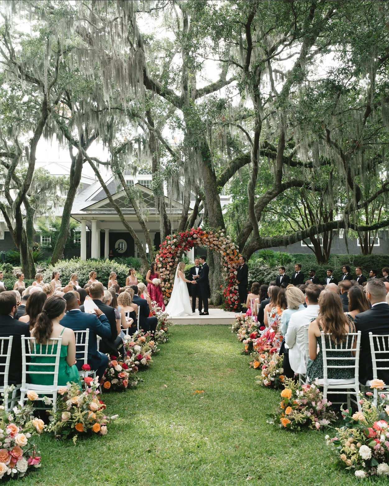 Outdoor wedding ceremony under sprawling live oaks with Spanish moss, featuring a colorful floral arch and a lush, flower-lined aisle with guests seated on both sides.