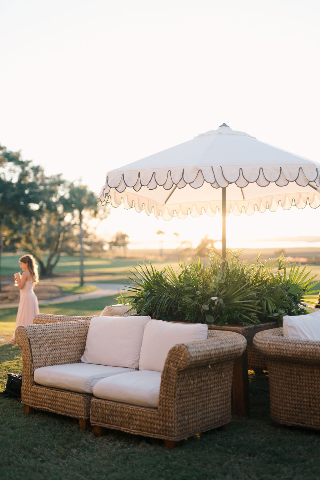 A cozy outdoor seating arrangement with wicker sofas and a scalloped umbrella, bathed in warm sunset light during a wedding celebration. A young girl in the background enhances the tranquil ambiance.
Anne Rhett Photography
Gathering Floral and Event