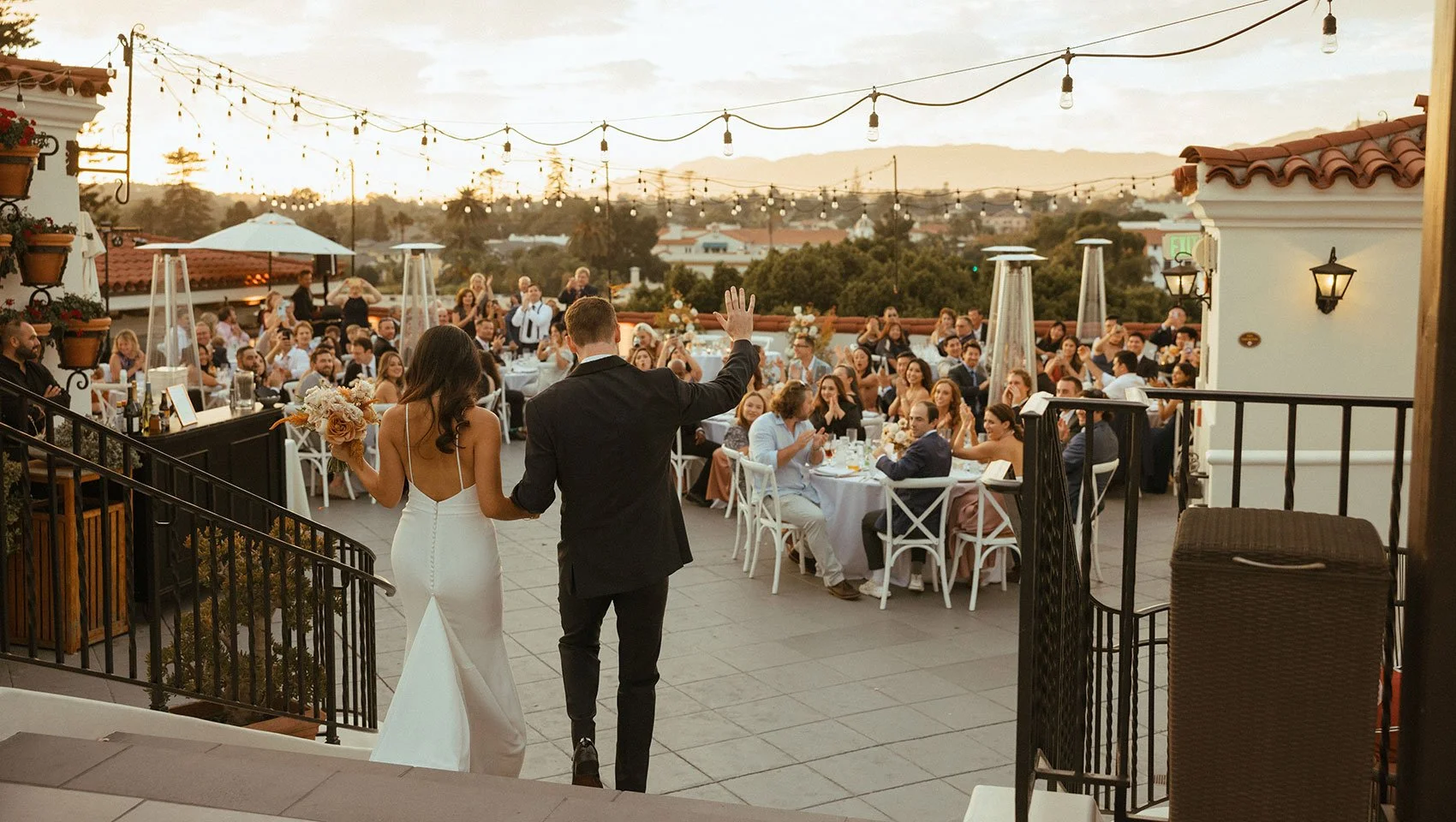 Bride and groom enter a rooftop wedding reception in Santa Barbara at sunset under string lights with mountain views.
