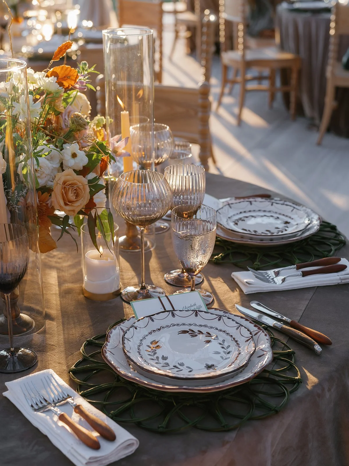 An intimate wedding tablescape with patterned china, woven green chargers, amber glassware, taper candles, and soft floral arrangements styled for a sophisticated reception.