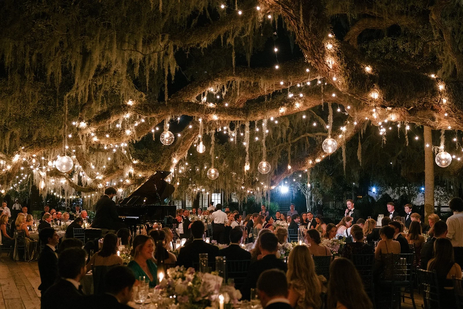 Outdoor Kiawah Island wedding reception beneath Spanish moss and warm string lights, with seated guests, candlelit tables, and a grand piano.