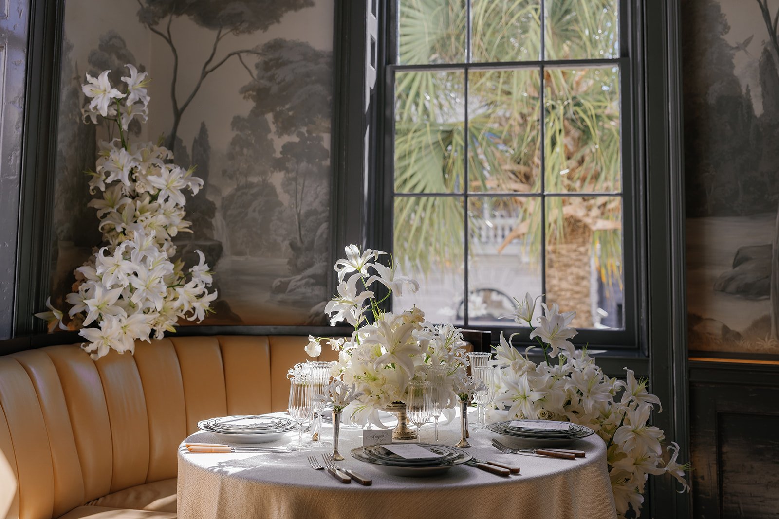 A round table dressed in ivory linen with black-and-white china, ribbed glassware, and abundant white lilies beside tall windows and mural wallpaper.