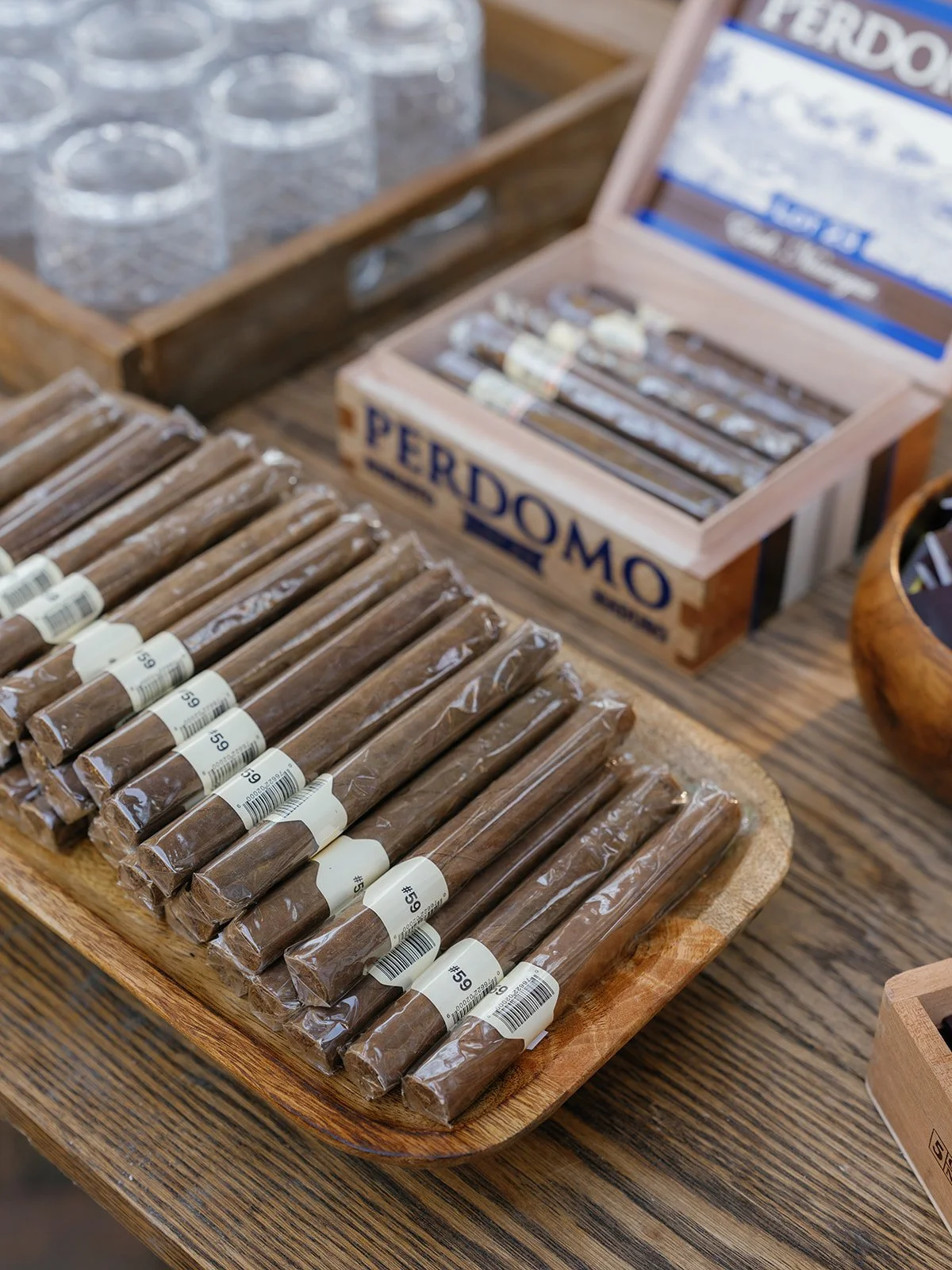 Cigar station with wrapped cigars arranged on a wooden tray beside open cigar boxes and glassware at a wedding welcome party.