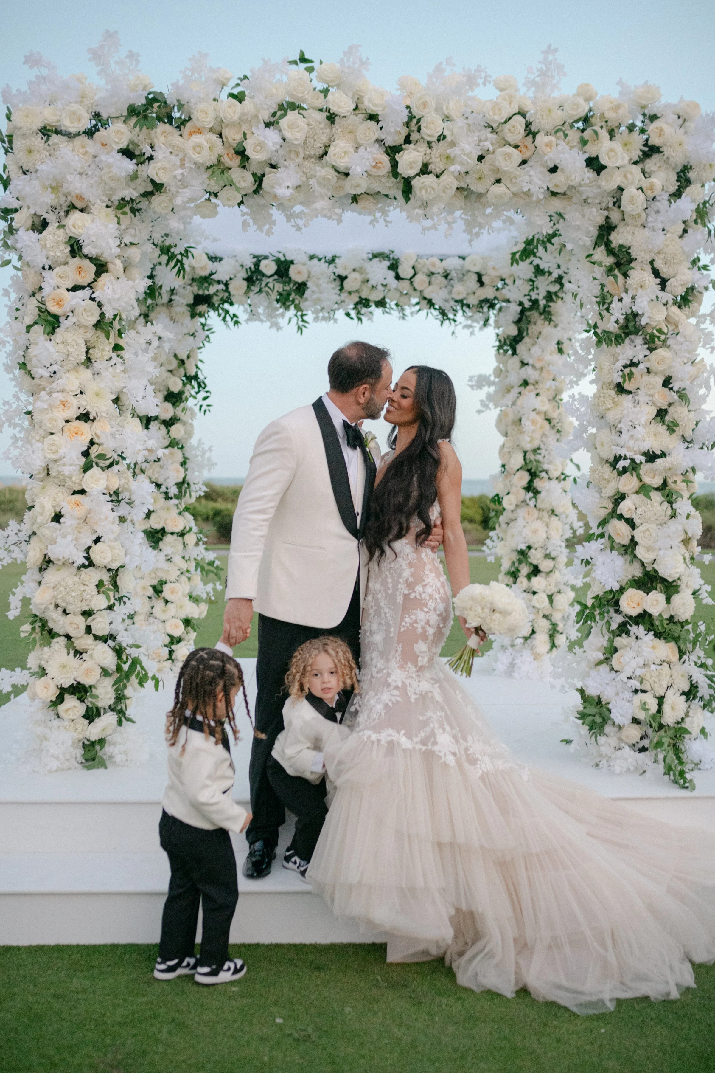 Newly married couple with two children beneath a large white floral ceremony arch at The Sanctuary on Kiawah Island.