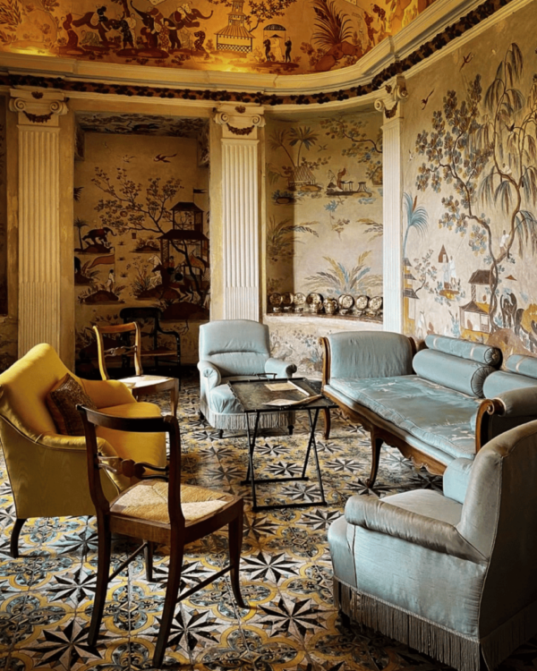 Painted salon inside a Tuscan villa with frescoed walls, patterned tile floors, and vintage seating in soft blue and gold tones.