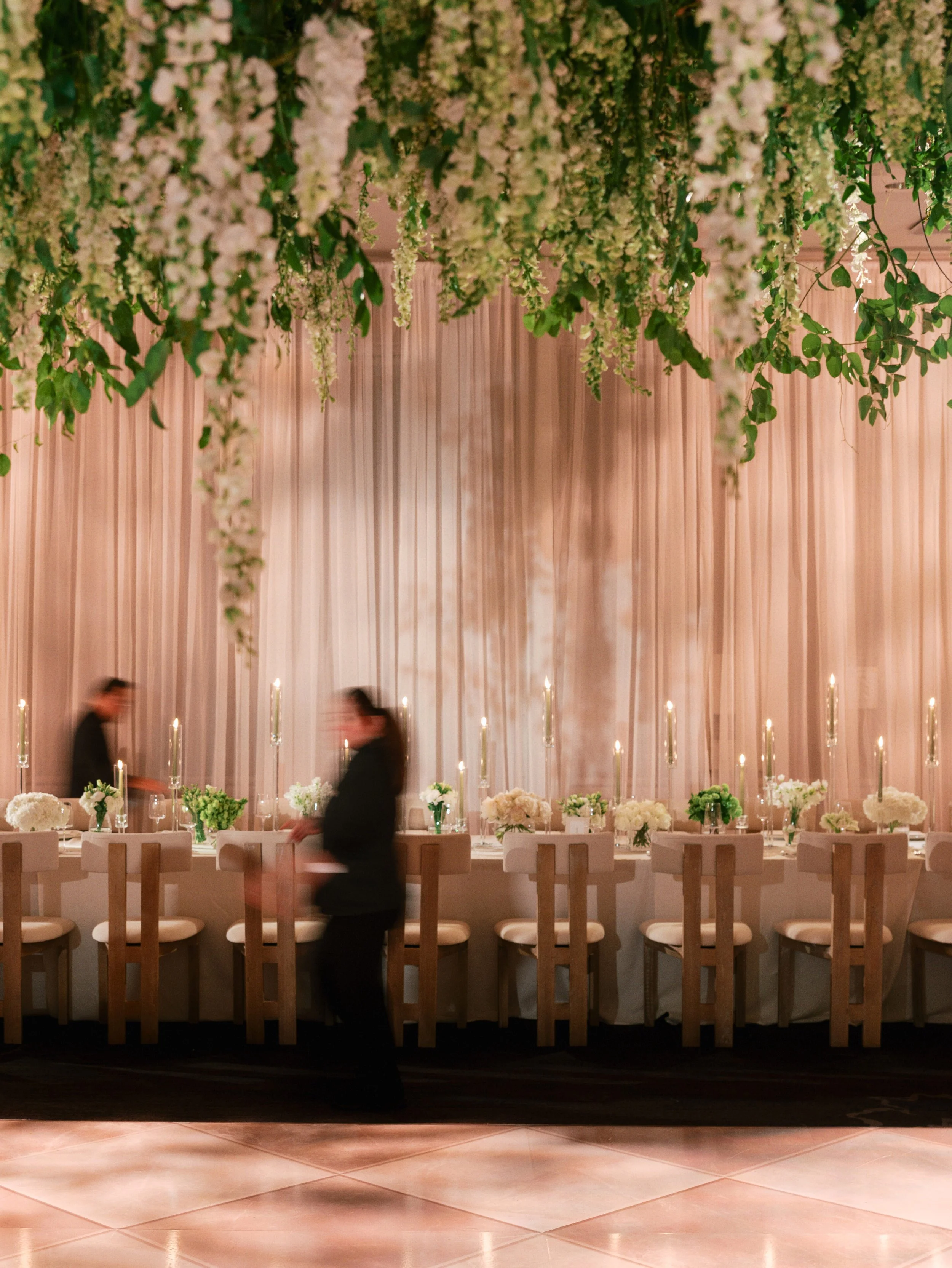 Candlelit reception with long tables and hanging greenery and white florals, with blurred staff moving through the room during setup or service.