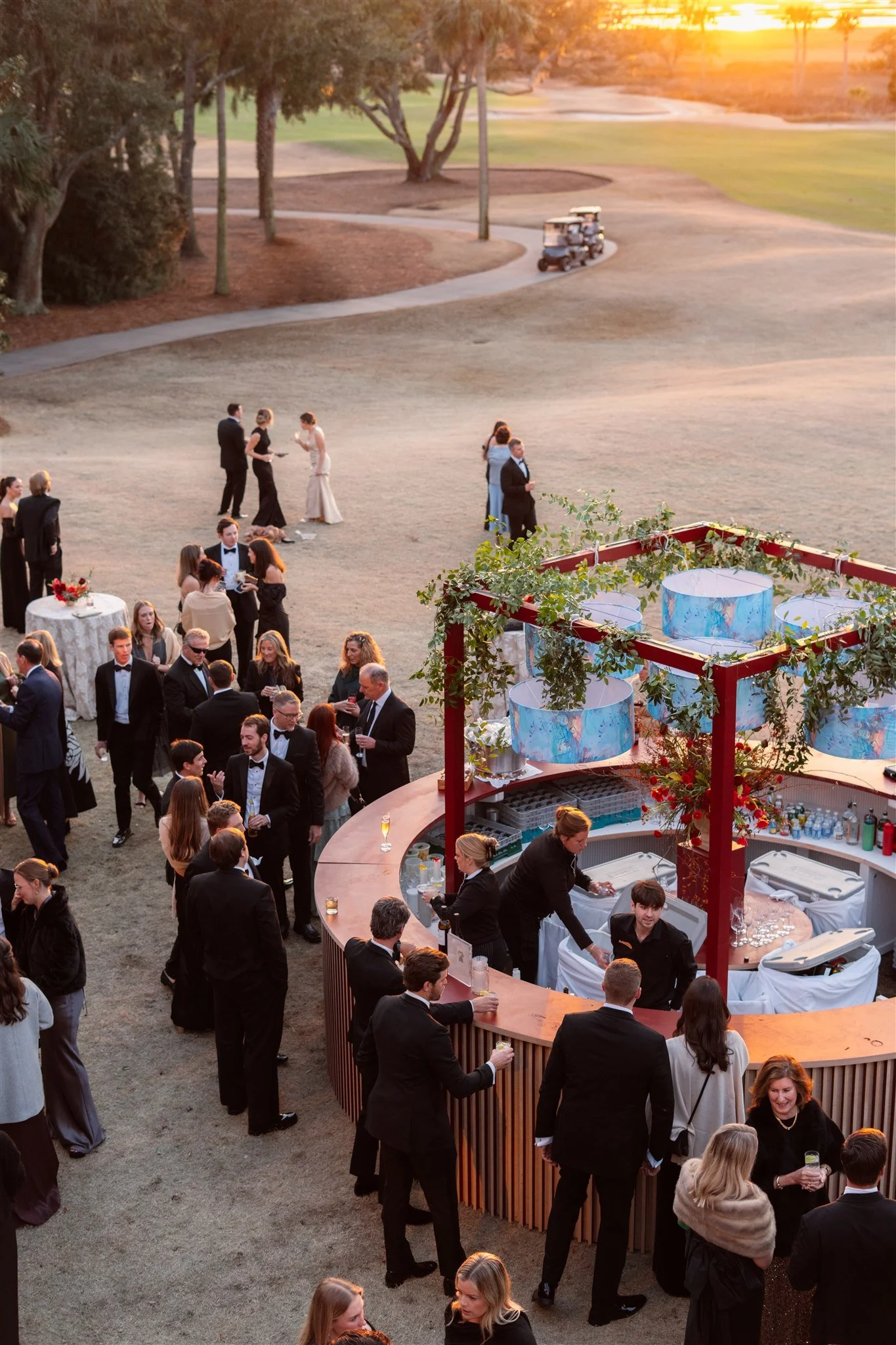 Overhead view of a black-tie wedding cocktail hour with guests gathered around a curved outdoor bar at sunset.