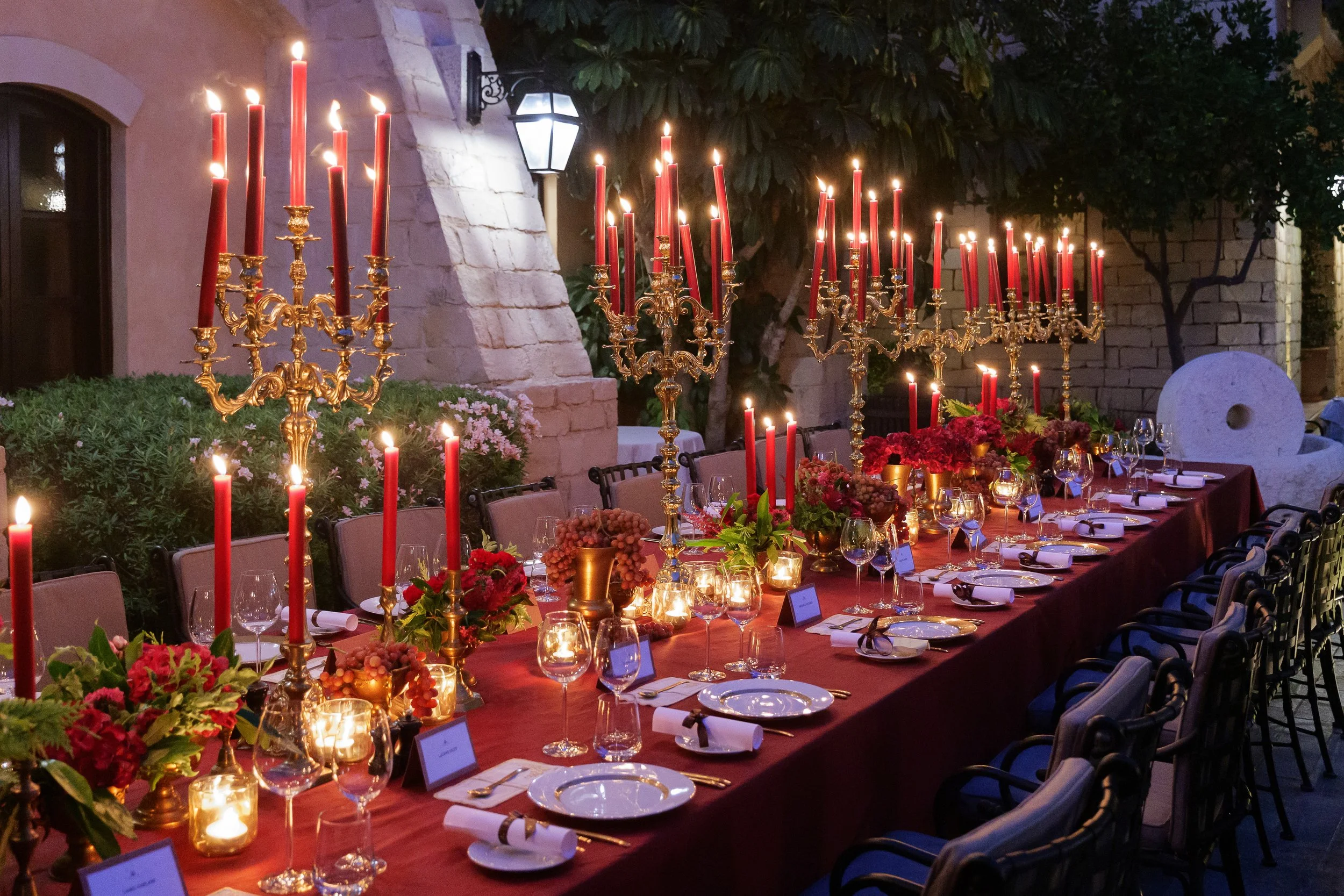 Candlelit courtyard dinner with a long red table, brass candelabras, red taper candles, and rich floral arrangements.