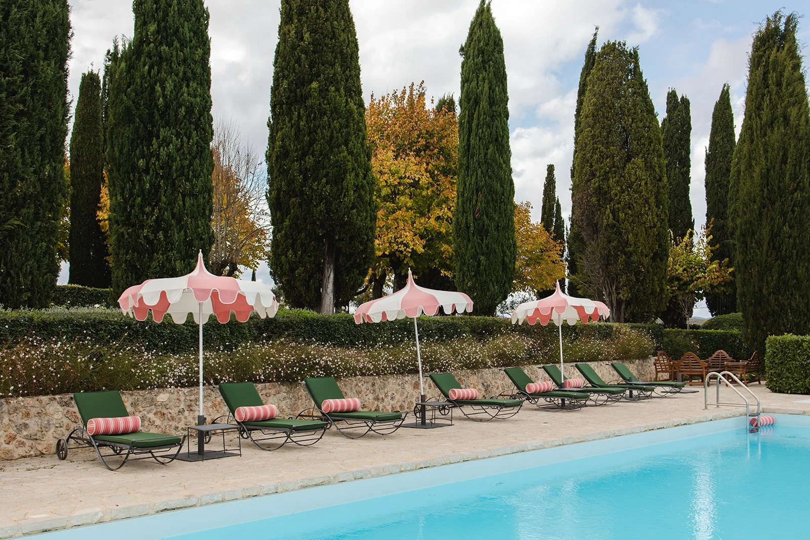 Poolside at a Tuscan villa with green loungers, pink scalloped umbrellas, stone decking, and tall cypress trees lining the property.