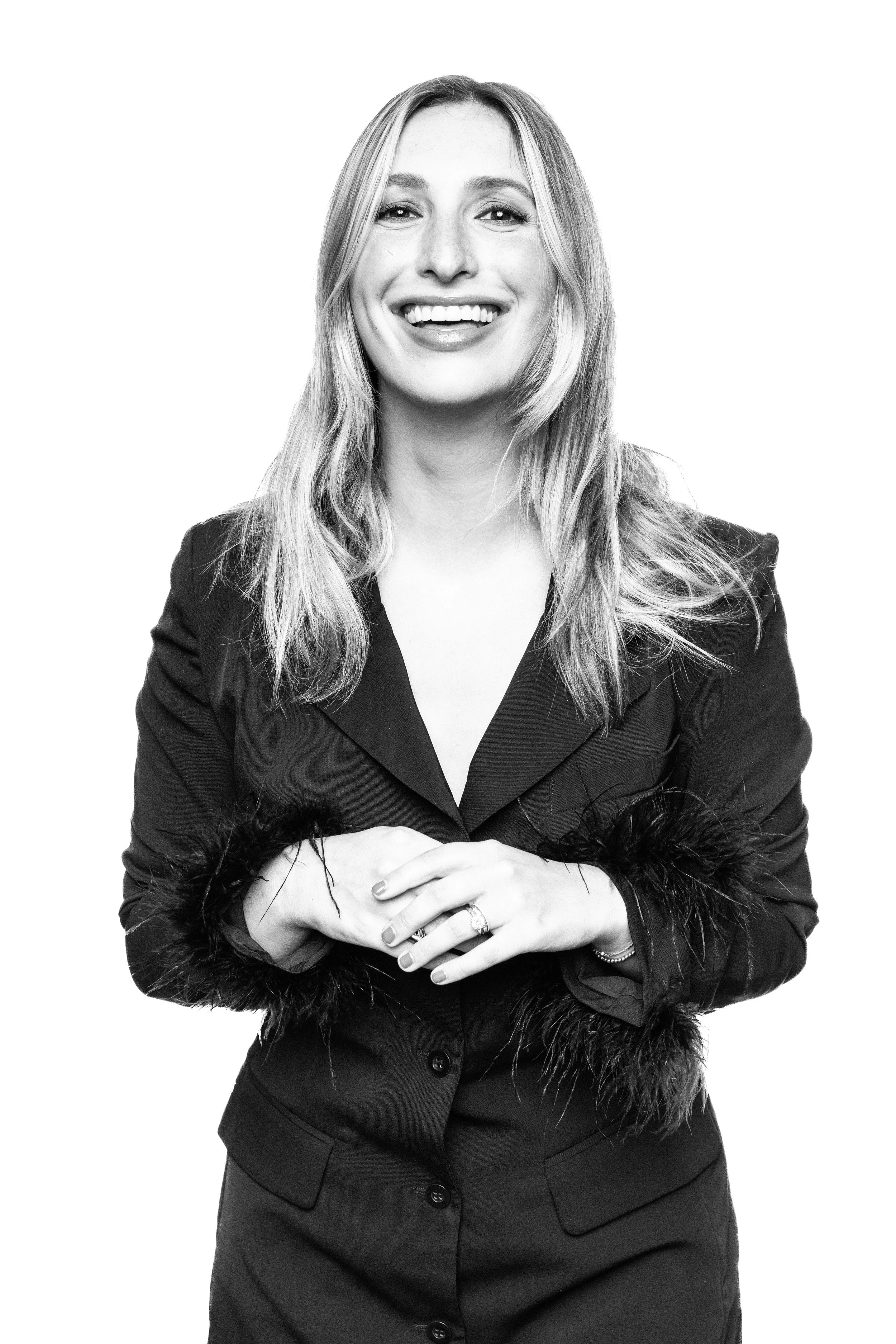 Black-and-white portrait of Sarah Schreiber smiling, wearing a tailored blazer with feathered cuffs against a white background.
